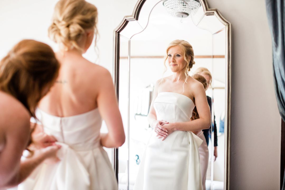 CJ's Off The Square | Bride looking at herself in the mirror while bridesmaid puts the final touch on her dress