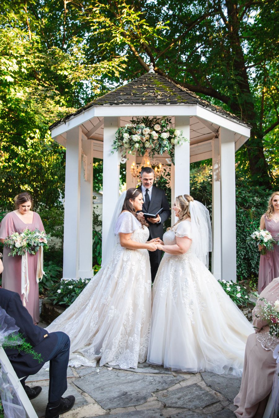 Brides holding hands in front of gazebo of garden wedding venue / Romantic / Summer / September / Pink / Dusty Rose / Cream